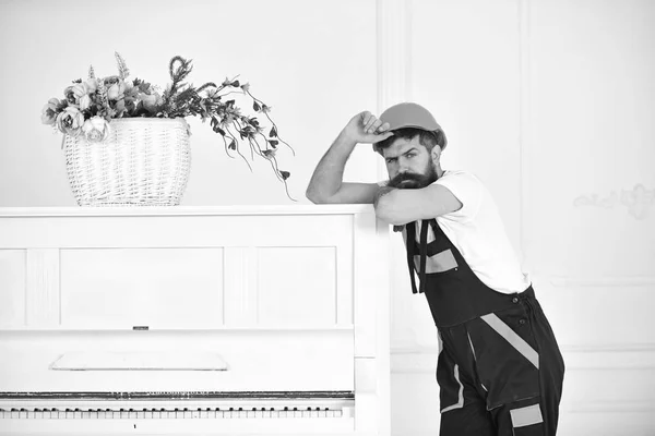 Tipo frunciendo el ceño con bigote elegante levantando la ceja. Mover apoyado en el piano blanco con pequeñas flores de color rosa y púrpura en la parte superior de ella. Hombre en casco naranja de pie aislado sobre fondo blanco — Foto de Stock