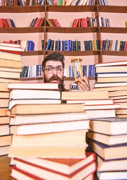 Czas przepływu koncepcja. Człowiek na zaskoczony twarz posiada klepsydra podczas studiów, regały na tle. Nauczyciel lub student z broda studia w bibliotece. Człowieku naukowiec w okularach wygląda poziomie klepsydry — Zdjęcie stockowe