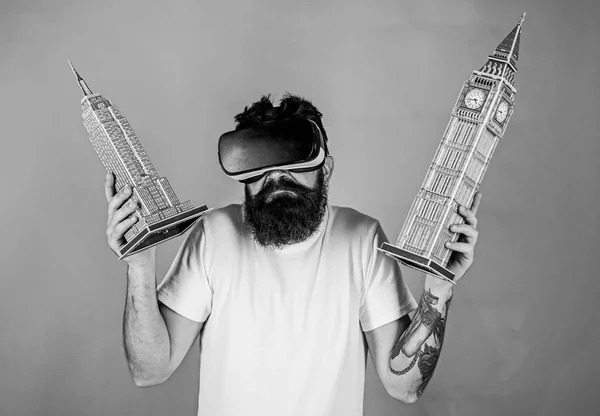 Man on confused face study architecture or design in virtual reality. Guy in VR glasses holds Big Ben and Empire State Building. 3D design concept. Man with beard in VR glasses, green background