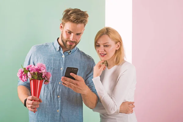 Guy with phone and bouquet of flowers, pastel pink and green background. Romantic concept. Man shows photo on smartphone to girl, sweet memories of their relations. Couple in love interested by phone