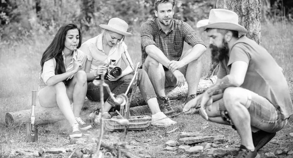 Amici gruppo turistico rilassante vicino al falò. Salsiccia arrosto Hipster mentre gli amici parlano condividere l'impressione e guardare le foto sulla macchina fotografica. Turisti che fanno merenda con arrosto sul cibo fuoco — Foto Stock