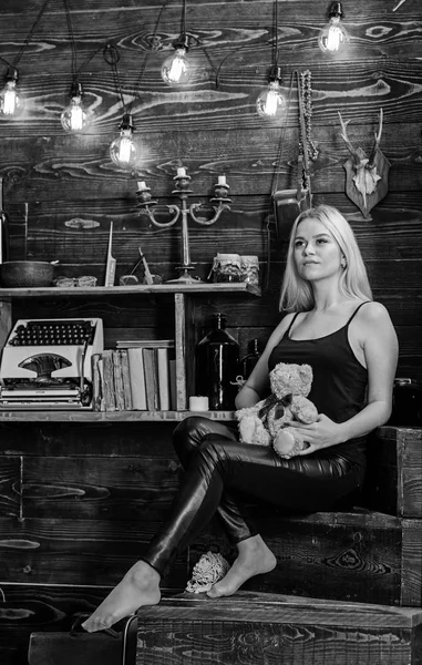 Girl in black clothes holds teddy bear toy in hand, wooden interior on background. Woman on dreamy face relaxing in wooden interior. Lady blonde enjoy leisure with teddy bear. Rest and relax concept