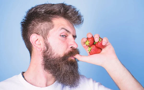 Man strict face enjoy fresh drink strawberry juice. Fresh juice concept. Man drinks strawberry juice suck thumb as drink straw blue background. Hipster bearded holds strawberries fist as juice bottle
