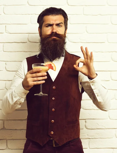 handsome bearded man with long beard and mustache has stylish hair on serious face holding glass of alcoholic beverage in vintage suede leather waistcoat on white brick wall studio background