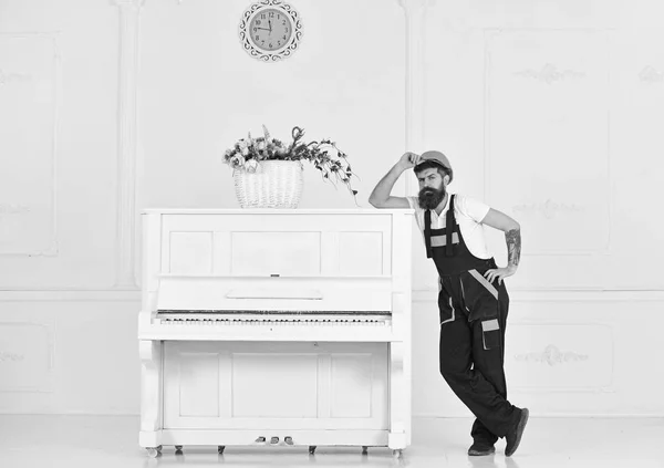 Hombre con barba, trabajador con mono y casco apoyado en el piano, fondo blanco. El cargador mueve el instrumento de piano. Courier entrega muebles en caso de mudanza, reubicación. Concepto de servicio de entrega — Foto de Stock