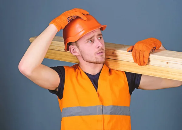 Uomo in casco, cappello rigido e guanti protettivi regge travi in legno, fondo grigio. Concetto di sicurezza e protezione. Falegname, falegname, forte costruttore su viso serio porta trave di legno sulla spalla — Foto Stock