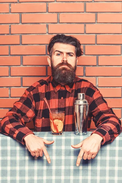 Bar menu concept. Bartender recommends to try beverage. Barman with beard on strict face pointing down with index finger. Man in checkered shirt near shaker and cocktail, brick wall background — Stock Photo, Image