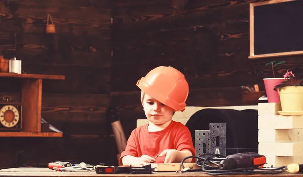 Jongen spelen als bouwer of hersteller, werken met hulpmiddelen. Jeugd concept. Kind droomt van toekomstige carrière in het platform of gebouw. Kid jongen in oranje hard hoed of helm, studie kamer achtergrond — Stockfoto