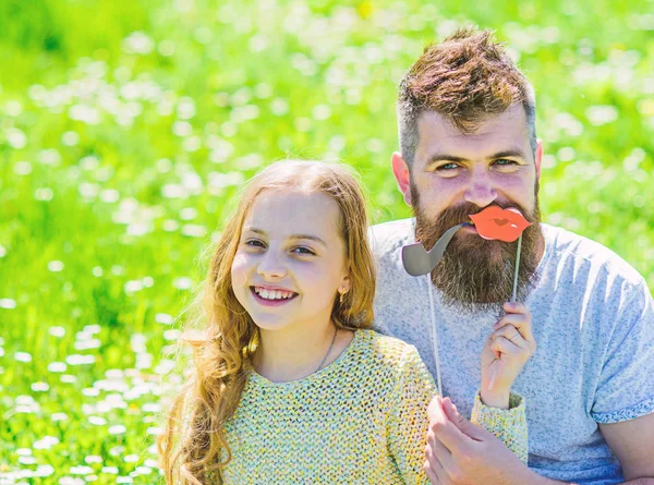 Family spend leisure outdoors. Dad and daughter sits on grass at grassplot, green background. Child and father posing with pipe and lips photo booth attributes. Fatherhood concept — Stock Photo, Image