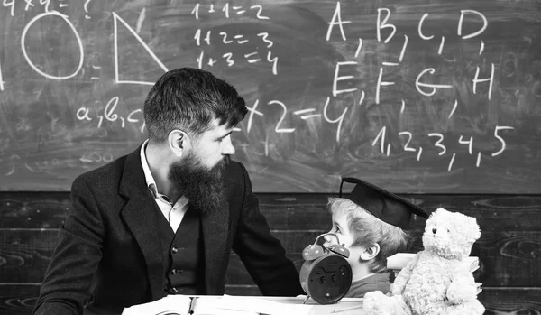 少年は、大学院のキャップの子見て落書き黒板に先生が説明しながら。ひげの先生は、父は、教室の背景に黒板に幼い息子を教えています。個人レッスンのコンセプト — ストック写真
