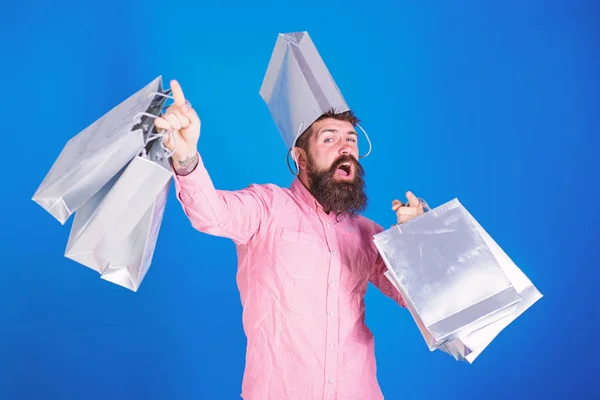 Hipster mit fröhlichem Gesicht und Tasche auf dem Kopf sind süchtig nach Shopaholic. Mann mit Bart und Schnurrbart trägt Einkaufstaschen, blauer Hintergrund. Einkaufskonzept. Mann kauft in der Verkaufssaison mit Rabatten ein — Stockfoto