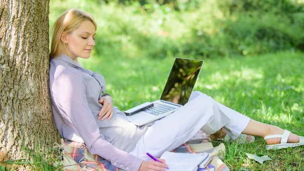 Образовательные технологии и интернет-концепция. Девушка, работающая с ноутбуком в парке, сидит на траве. Природный офис. Преимущества работы на открытом воздухе. Женщина с ноутбуком работает на открытом воздухе опираясь на ствол дерева — стоковое фото