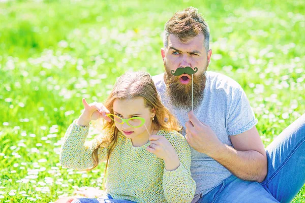 Father posing with mustache and child posing with eyeglasses photo booth attribute. Detectives concept. Dad and daughter sits on grassplot, grass on background. Family spend leisure outdoors — Stock Photo, Image