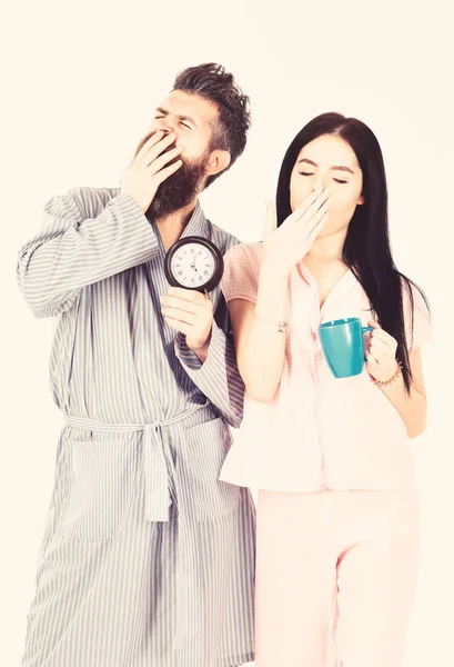 Pareja enamorada, familia joven en pijama, albornoz aislado sobre fondo blanco. Concepto matutino perfecto. Una pareja se despertó temprano en la mañana. Chica con taza de café, el hombre tiene el reloj en la mano y bostezar —  Fotos de Stock