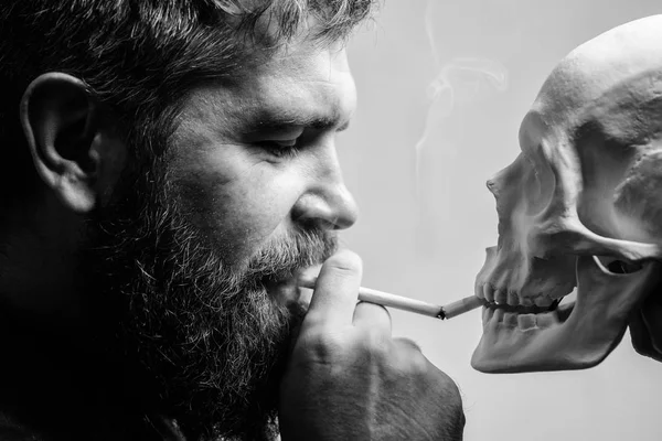 Man smoking cigarette near human skull symbol death. Nicotine destroy health. Harmful habits. Smoking is harmful. Habit to smoke tobacco bring harm to your body. Smoking cause health damage and death