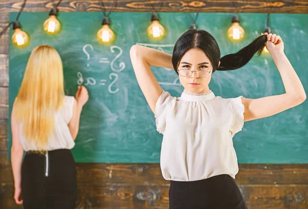 Studenten en stagiairs concept. Aantrekkelijke vrouwen voorbereiding voor les. Student, leraar in brillen staan in de klas. Meisje ziet er aantrekkelijk uit, terwijl dame schrijven op schoolbord achtergrond, defocuse — Stockfoto