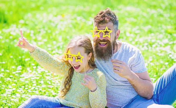 Star and fame concept. Dad and daughter sits on grass at grassplot, green background. Child and father posing with star shaped eyeglases photo booth attribute at meadow. Family spend leisure outdoors — Stock Photo, Image