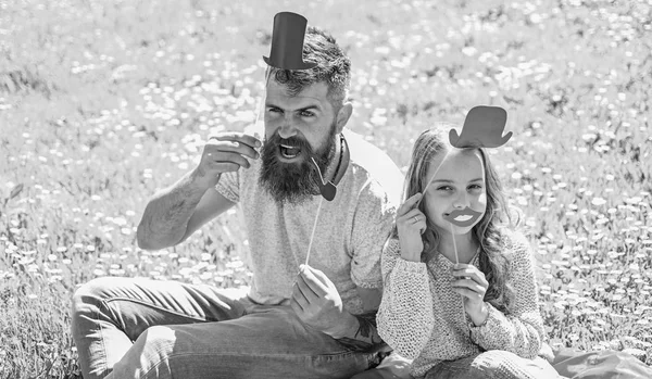 Aristocrates concept. Dad and daughter sits on grass at grassplot, green background. Family spend leisure outdoors. Child and father posing with top hat, lips and pipe photo booth attributes