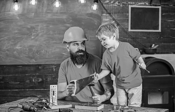 父は、学校のワーク ショップでさまざまなツールを使用するほとんどの息子を教える保護用のヘルメットでひげを持つ親。父権のコンセプトです。少年は、おもちゃで遊ぶ元気な子供を見て、お父さんと使用ツールを学習 — ストック写真