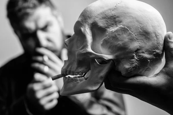 Man smoking cigarette near human skull symbol of death. Harmful habits. Smoking cause health damage and death. Destroy your health. Smoking is harmful. Habit to smoke tobacco bring harm to your body
