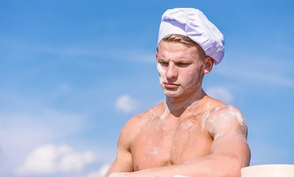 Chef en sombrero blanco en cara estricta, cielo en el fondo. Concepto de chef sexy. Hombre con apariencia atractiva trabaja como cocinero o panadero, espacio de copia. Hombre con torso muscular cubierto de harina se ve atractivo — Foto de Stock