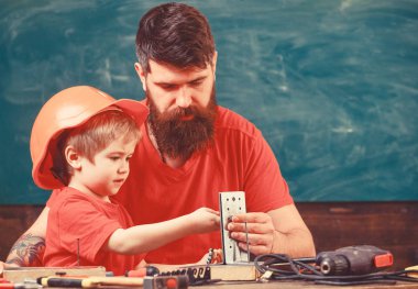 Mens work concept. Father with beard teaching little son to use tools in classroom, chalkboard on background. Boy, child busy in protective helmet makes by hand, repairing, does crafts with dad clipart