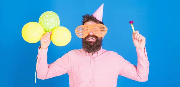 Cintura para cima tiro de boa aparência homem positivo com barba segurando balões de hélio e assobio sorrindo amplamente, se divertindo na festa. Pessoas, alegria, entretenimento e celebração conceito — Fotografia de Stock