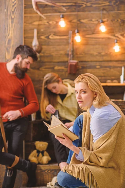 Mujer con cabello rubio y aspecto astuto leyendo novela favorita. Esposa de leñador disfrutando de un nuevo libro. Hombre barbudo afilando cuchillo y charlando con su hija. Vacaciones en zona rural, concepto fuera de la ciudad —  Fotos de Stock