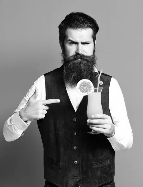 Bel homme barbu avec longue barbe et moustache a des cheveux élégants sur le visage sérieux tenant verre de cocktail alcoolisé en cuir suédé vintage gilet sur fond de studio rouge — Photo