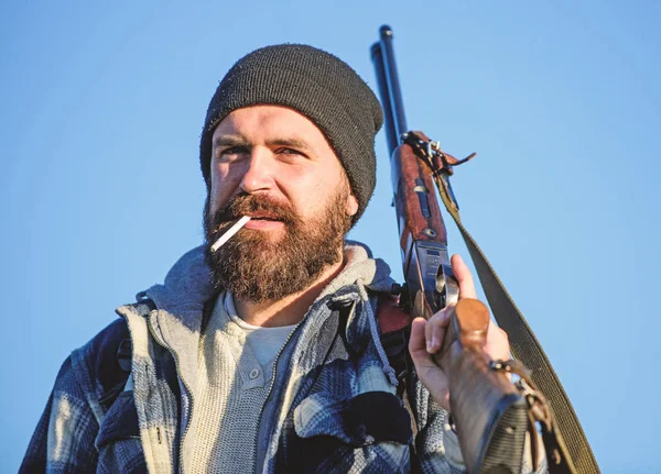 Guy bearded hunter spend leisure hunting and smoking. Hunting masculine hobby concept. Man brutal bearded guy gamekeeper blue sky background. Brutality and masculinity. Hunter with rifle gun close up