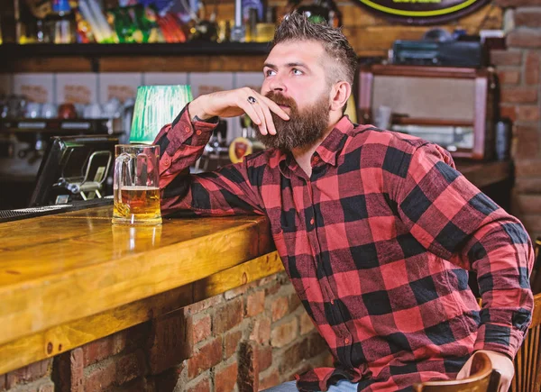 Hipster relaxing at bar with beer. Order alcohol drink. Bar is relaxing place have drink and relax. Man with beard spend leisure in dark bar. Brutal hipster bearded man sit at bar counter drink beer