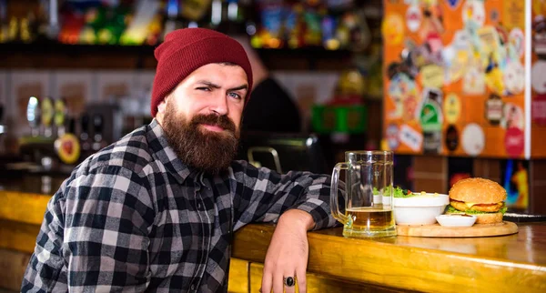 Hipster relaxing at pub. Pub is relaxing place to have drink and relax. Man with beard drink beer eat burger menu. Enjoy meal in pub. High calorie snack. Brutal hipster bearded man sit at bar counter