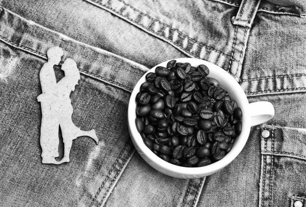 Керамическая чашка с кофейными зёрнами и деревянный силуэт танцующей пары на фоне джинсов. Романтический перерыв на кофе. Кружка кофе в зернах на белом фоне. Наслаждайся кофе. Дата в концепции кафе — стоковое фото
