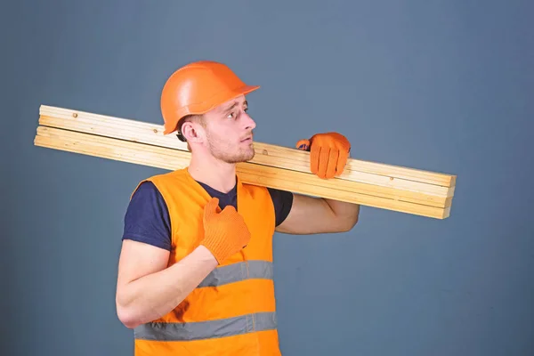 Wooden materials concept. Carpenter, woodworker, strong builder on serious face carries wooden beam on shoulder. Man in helmet, hard hat and protective gloves holds wooden beam, grey background — Stock Photo, Image