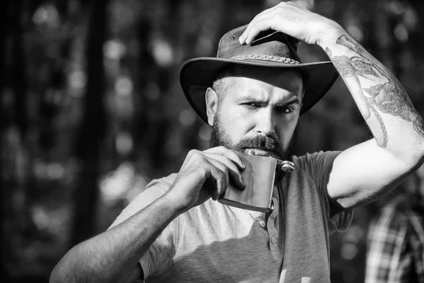 L'aventure commence dès la première gorgée. Boisson alcoolisée. L'homme nomade porte de l'alcool avec lui. Un cow-boy barbu et brutal boit de l'alcool. Hipster avec barbe boire alcool nature fond déconcentré — Photo