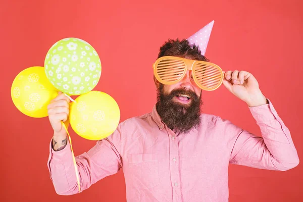 Hipster gucken aus einer riesigen Sonnenbrille. Mann mit Bart und Schnurrbart auf glücklichem Gesicht hält Bündel Luftballons, roter Hintergrund. Festkonzept. Mann mit Partyhut und Luftballons feiert — Stockfoto