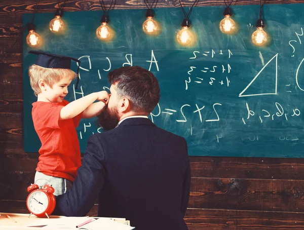 Guy vände tillbaka och små barn i klassrummet. Söt pojke leker med mustasch av mannen. Ler barn i examen cap sidovy — Stockfoto