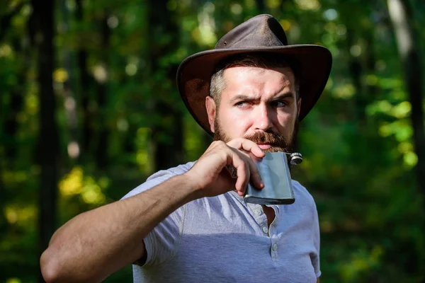 Hombre hipster relajarse en el bosque de otoño. Primavera tiempo soleado. camping y senderismo. macho maduro con aspecto brutal beber alcohol de matraz metálico. Barbudo hombre en vaquero sombrero caminar en parque al aire libre —  Fotos de Stock