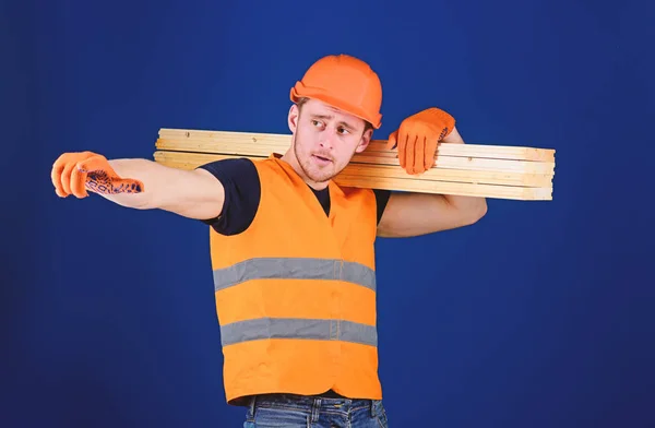 Tischler, Holzarbeiter, kräftiger Bauarbeiter auf dem Gesicht trägt Holzbalken auf der Schulter. Holzwerkstoffkonzept. Mann mit Helm, Schirmmütze und Schutzhandschuhen weist Richtung, blauer Hintergrund — Stockfoto