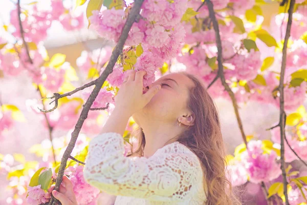 Menina no rosto sorridente em pé perto de flores sakura, desfocado. Conceito de perfume e fragrância. A criança bonita gosta do aroma de sakura no dia de primavera. Menina com cabelos longos ao ar livre, flor de cereja no fundo — Fotografia de Stock