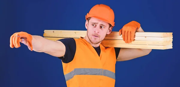 Ο άνθρωπος στο κράνος, κράνος και προστατευτικά γάντια που δείχνει την κατεύθυνση, μπλε φόντο. Ξύλινα υλικά έννοια. Ξυλουργός, ξυλουργός, ισχυρό οικοδόμος στο στοχαστικό πρόσωπο ασκεί ξύλινα δοκάρια στον ώμο — Φωτογραφία Αρχείου