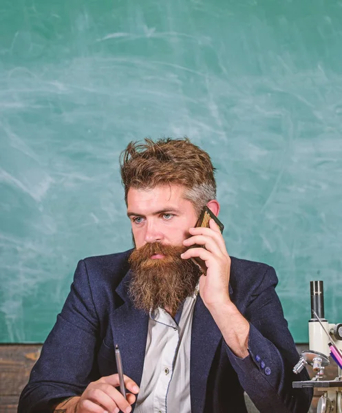 Keep in touch. School teacher call mobile phone while sit classroom chalkboard background. Pedagogue keep in touch with colleagues. Teacher bearded man talk mobile phone. Call colleague ask advice