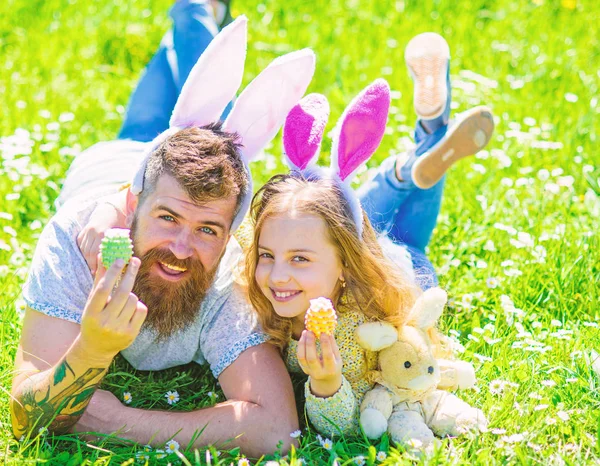 Koncepce rodinné dovolené. Otec a dcera na šťastné tváře pořádá velikonoční vajíčka, tráva na pozadí. Dívka s tátou ležící na trávě na slunečný den. Dítěte a muž s roztomilý zajíček uši hrát spolu — Stock fotografie