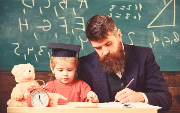 Busy kid studying at school. Teacher, father checking homework, helps to boy, son. Teacher in formal wear and pupil in mortarboard in classroom, chalkboard on background. Individual studying concept
