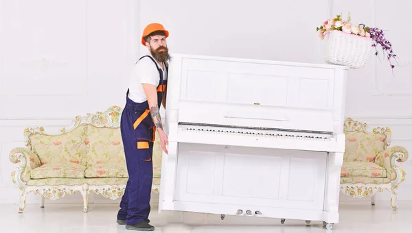 Loader moves piano instrument. Man with beard worker in helmet and overalls lifts up, efforts to move piano, white background. Heavy loads concept. Courier delivers furniture, move out, relocation