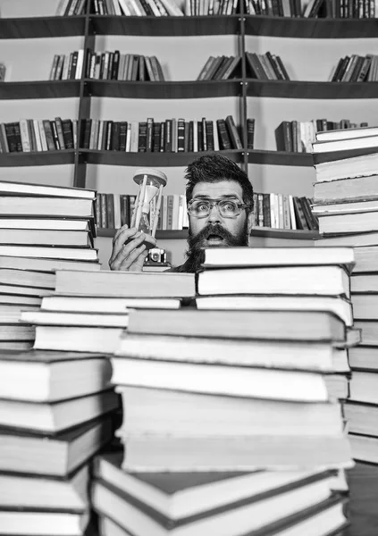 Conceito de fluxo de tempo. Homem no rosto surpreso detém ampulheta enquanto estuda, estantes de livros no fundo. Professor ou estudante com barba estudando na biblioteca. Homem, cientista de óculos olha para ampulheta — Fotografia de Stock