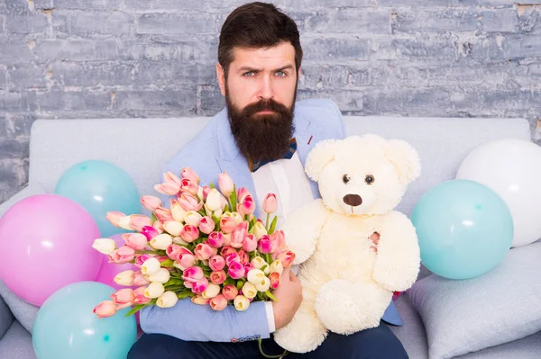 Macho getting ready romantic date. Man wear blue tuxedo bow tie hold flowers bouquet. Best boyfriend ever. Romantic man with flowers and teddy bear sit on couch waiting girlfriend. Romantic gift