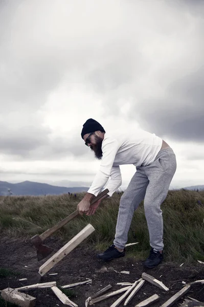 Ο άνθρωπος γένια hipster μπριζόλα ξύλο με τσεκούρι σε ορεινό τοπίο — Φωτογραφία Αρχείου