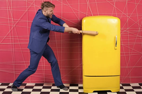 Man formal elegant suit beat with wooden bat retro vintage yellow refrigerator. Bachelor hungry want eat near fridge. Bright fridge household appliances. Hunger and appetite. Hungry attack concept