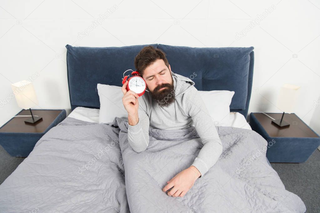 Need more sleep. Get up with alarm clock. Tips for waking up early. Man bearded sleepy face bed with alarm clock in bed. What terrible noise. Turn off that ringing. Problem early morning awakening